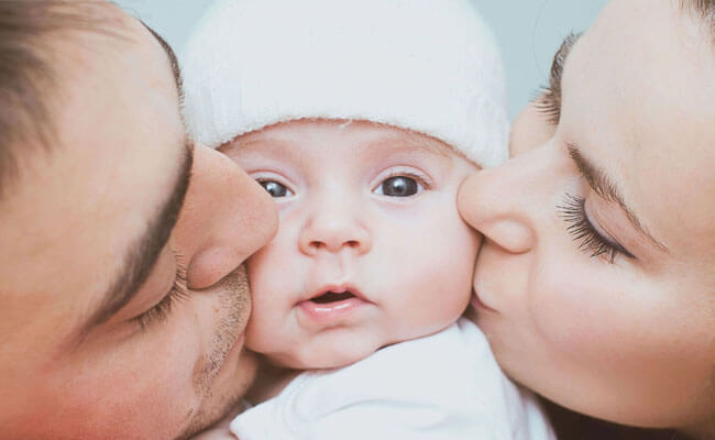 Picture of a young couple kissing the cheeks of their baby.  The picture is a close-up, with the baby looking directly at the camera.