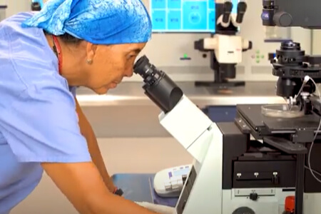 Picture of a laboratory technician looking through a microscope in an IVF fertility center.  The person is wearing gloves, a blue head covering and a blue smock.