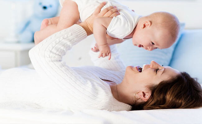 Picture of a happy mother playing with her baby. The woman is lying down, looking upward, smiling, and holding her baby above her.