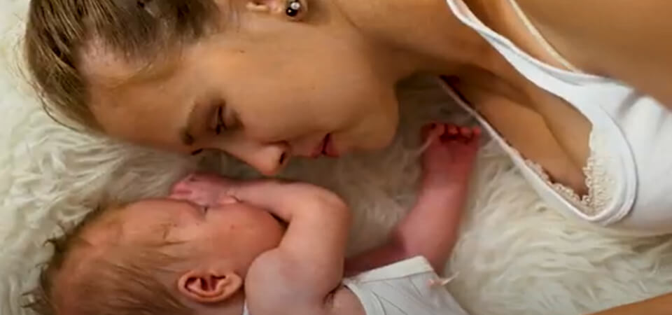 Picture of a happy mother looking at her sleeping baby.  The mother and baby are laying on their side, in bed.